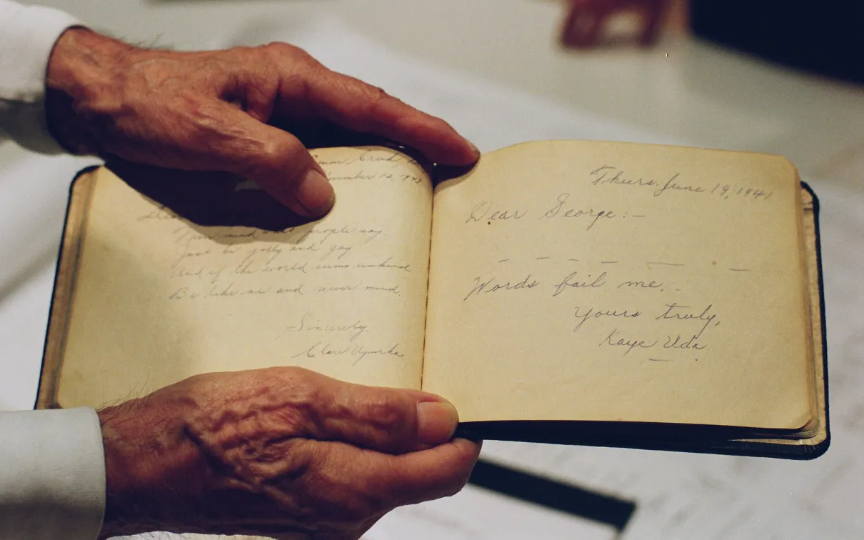 Photo of a pair of hands holding a handwritten book. The entry reads: Dear George, Words fail me. Yours truly, Kaye Uda.
