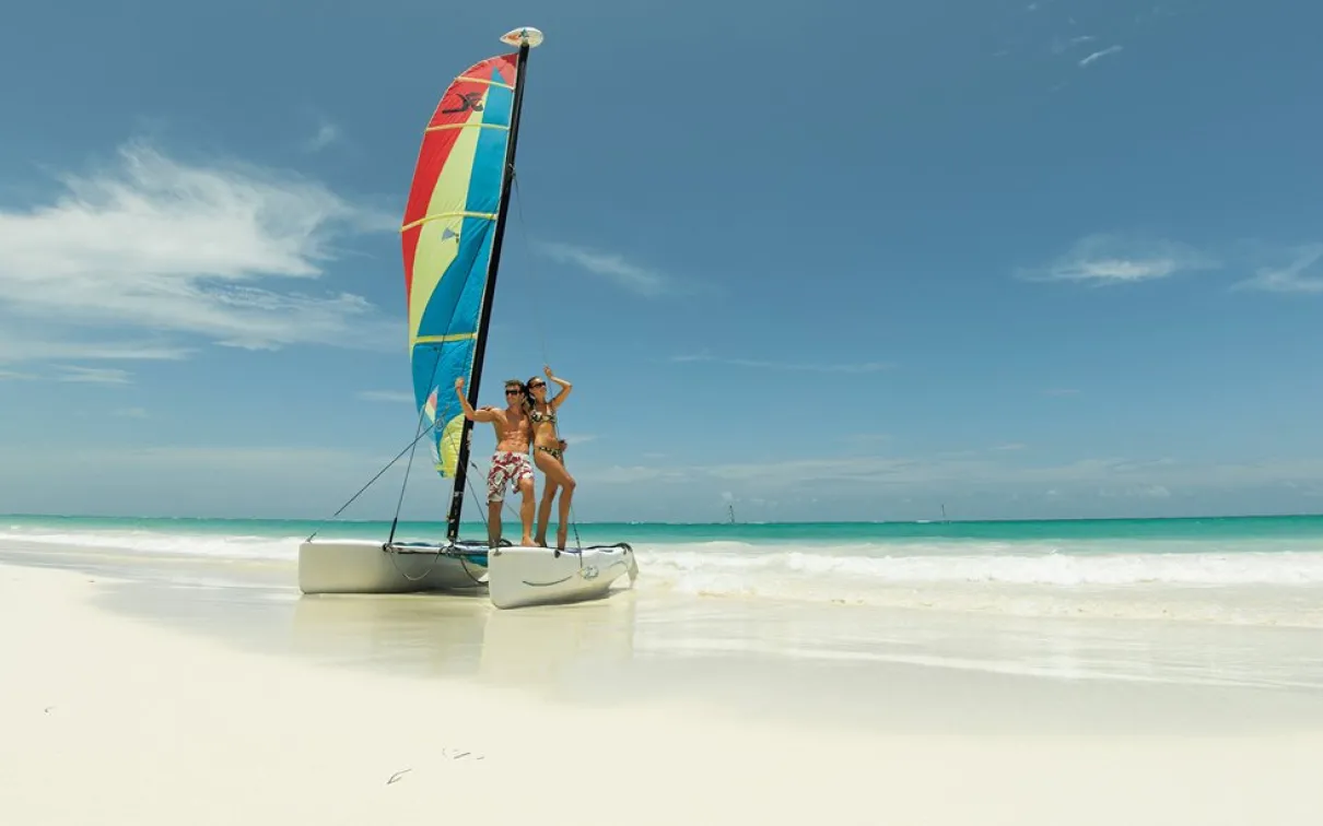 A man & a woman in bathing suits pose on a small catamaran on a sun-lit, white sand beach
