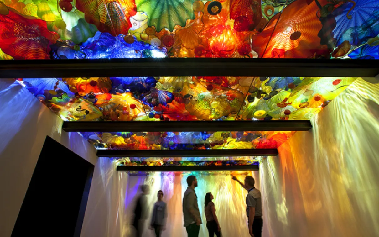 Visiteurs de l'exposition avec des sculptures en verre exposées au-dessus d'un plafond en verre rétro-éclairé.