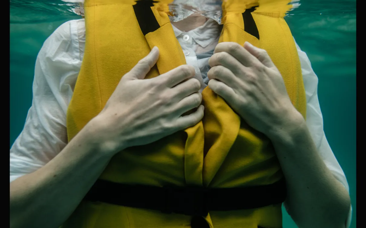 Image prise sous l'eau du torse d'une femme vêtue d'une chemise blanche et d'un gilet de sauvetage jaune.
