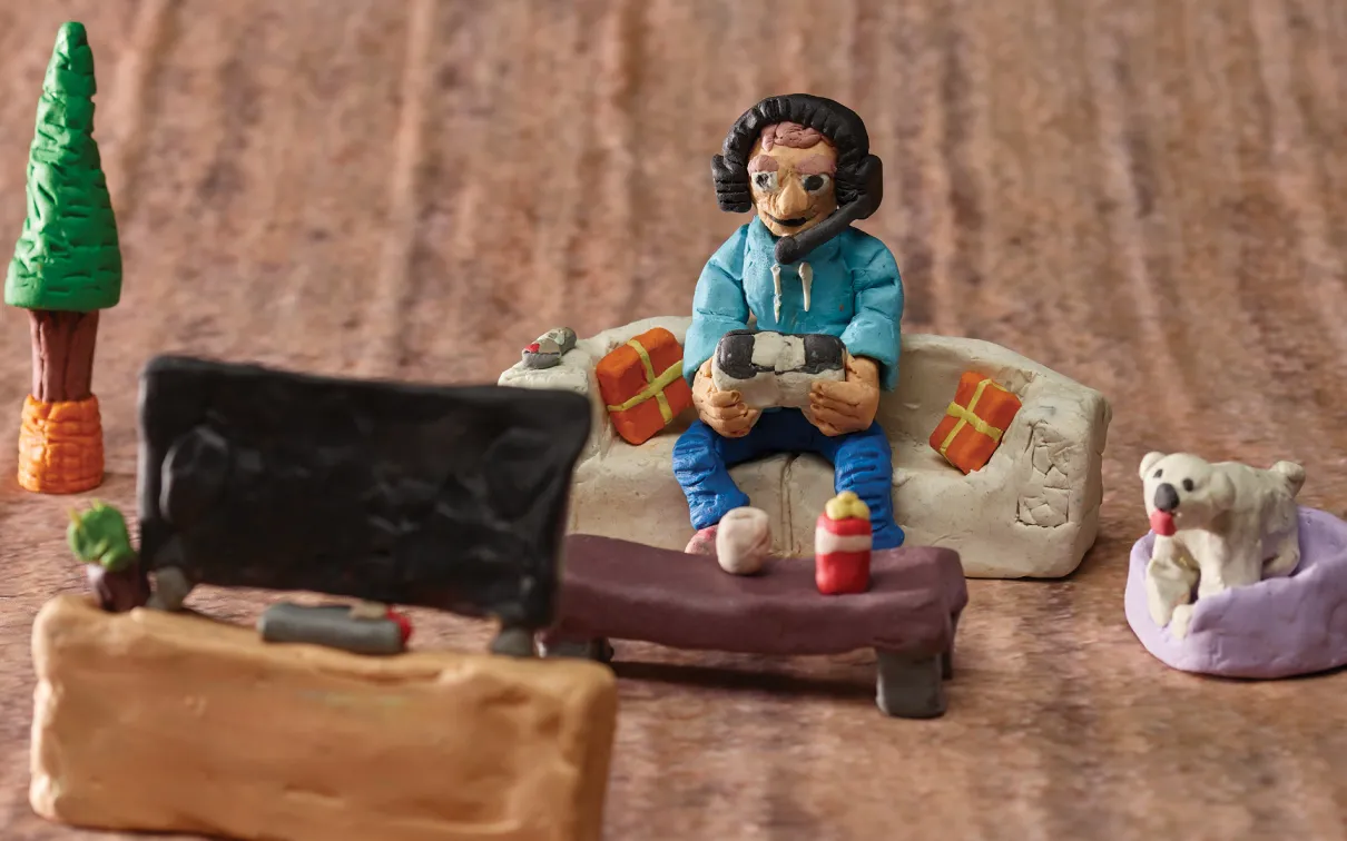Détail d'un diorama montrant une personne portant des écouteurs sur un canapé, jouant à un jeu vidéo tout en regardant un écran de télévision.