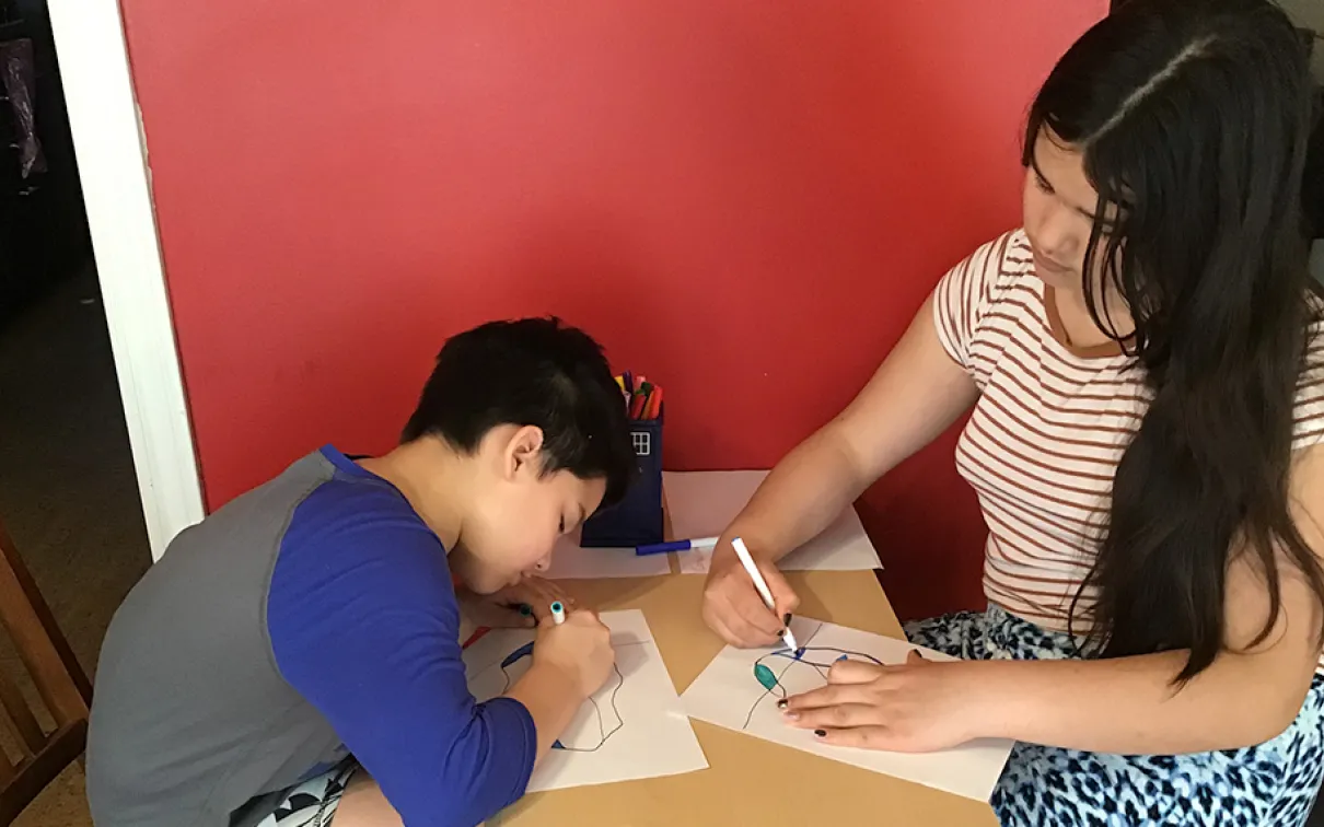 wo girls, one with short dark hair and the other with long dark hair, sit at a table and colour iceberg drawings with markers.
