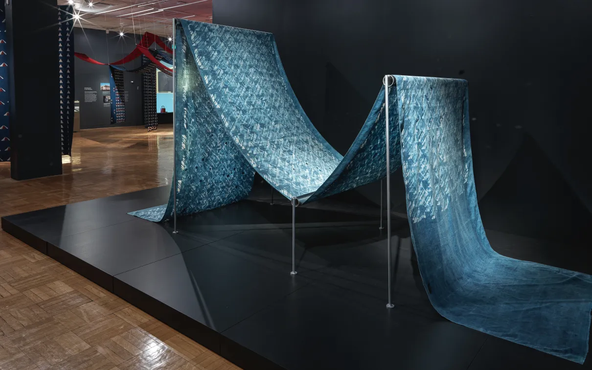 View of a length of dyed blue cotton fabric with mirror embellishments arranged on a frame to resemble fabric on a weaving loom in the exhibition Swapnaa Tamhane: Mobile Palace, at ROM.