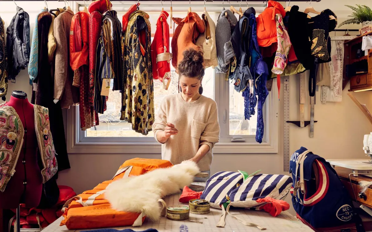 Image of the artist working at a large table containing works in progress, with a selection of lifejackets suspended along the wall behind.

