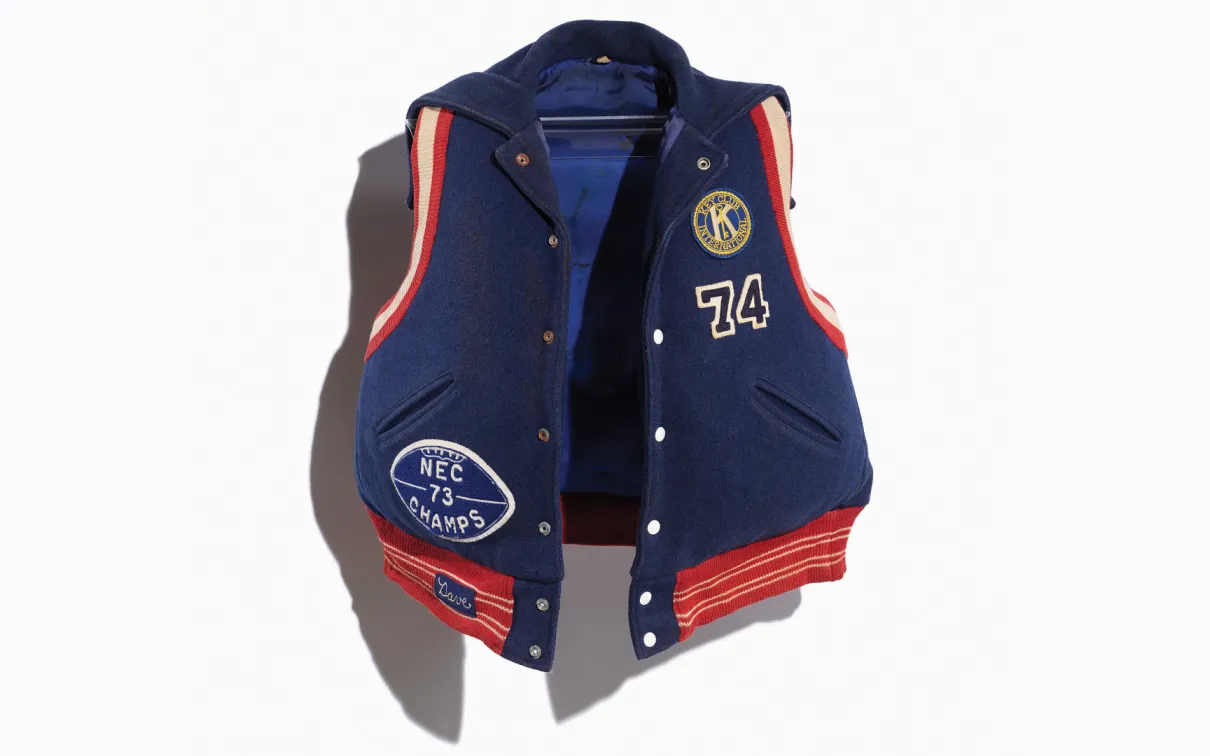 Image of a repurposed life jacket covered in a navy varsity jacket with orange and red trim and school letters and team patches/numbers. Image taken on a white background.

