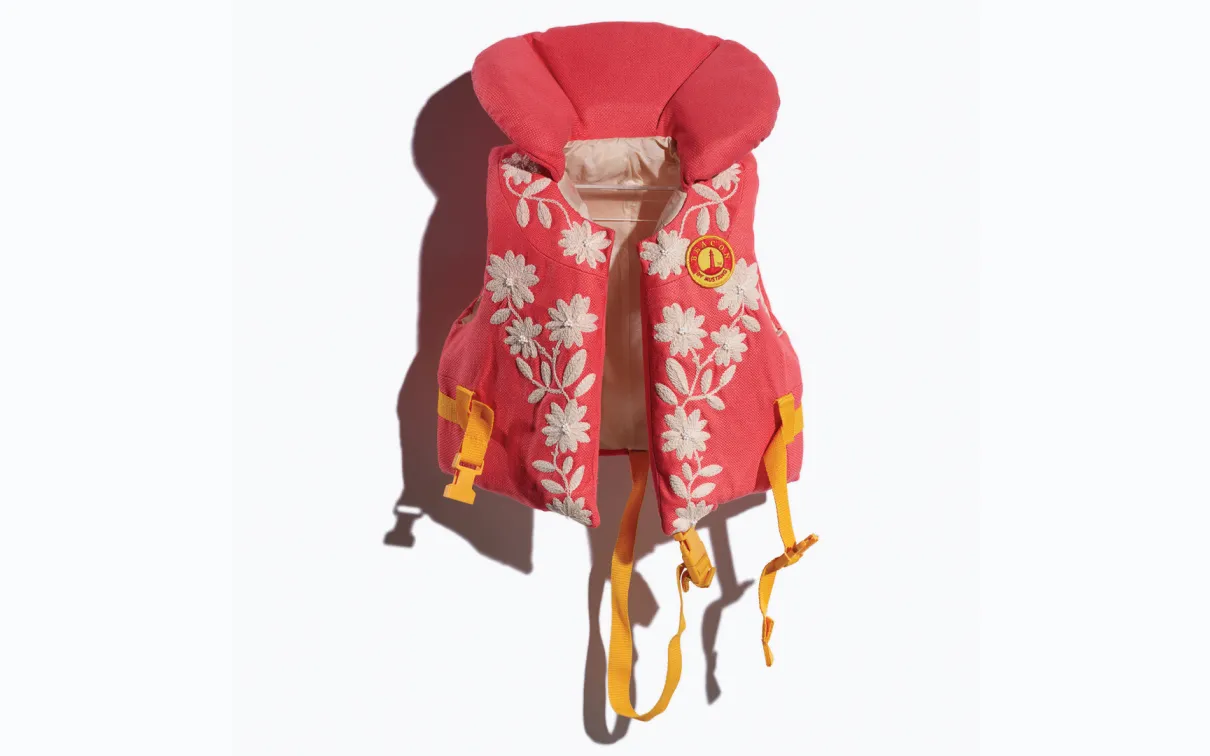 Image of a repurposed life jacket, covered in orange fabric covered with embroidery depicting white flowers. Image taken on a white background.
