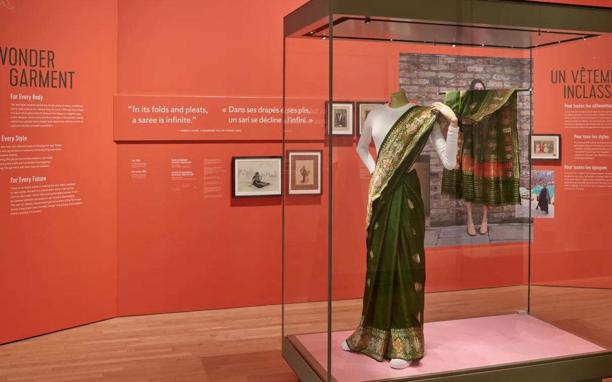 Installation présentant des panneaux de texte rouges derrière une vitrine contenant un sari de soie verte avec des fils métalliques tissés, porté sur un T-shirt blanc, sur un mannequin portant des chaussures de course blanches.