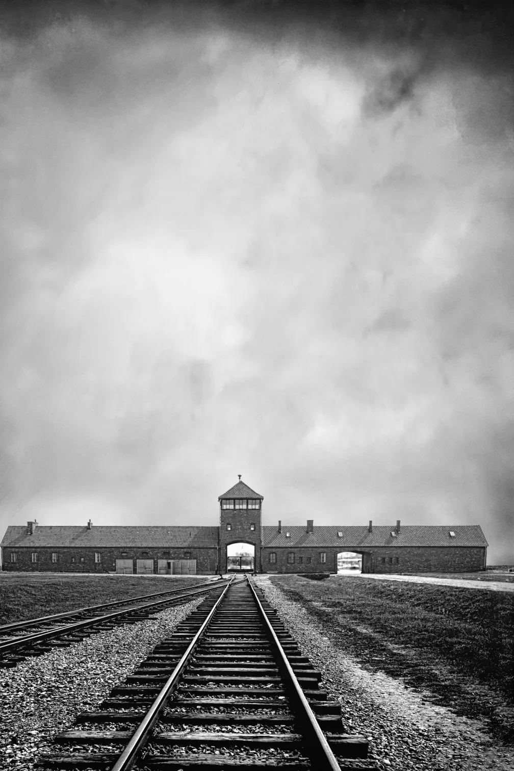 Black and white archival image showing train tracks in the foreground leading into the distance towards the entrance to the Auschwitz camp.