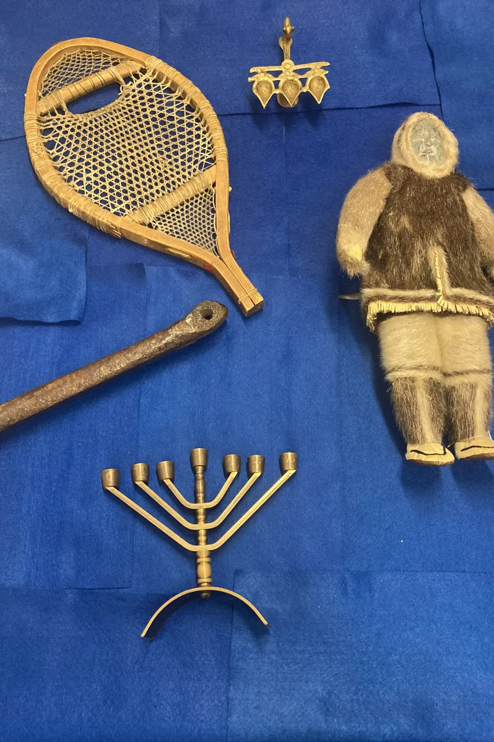 A selection of objects on a blue background.  The objects include an Inuit stone and sealskin doll, a Hannukiah, a medieval bone skate, a snowshoe, and a diya lamp.
