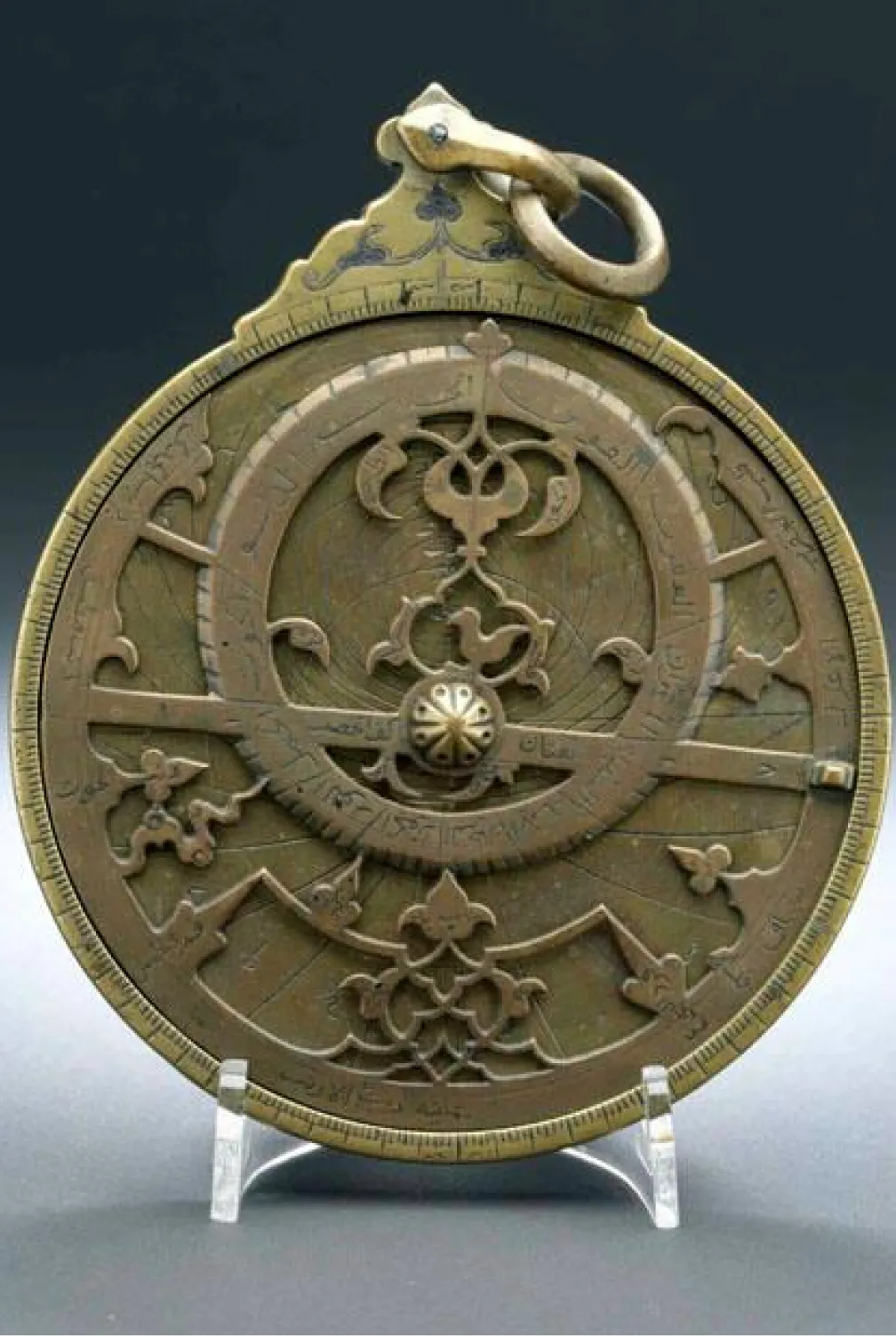 A bronze-coloured disk with carved details including an off-centre ring, leaves, and a small bird. There is a ring at the top similar to a pendant.
