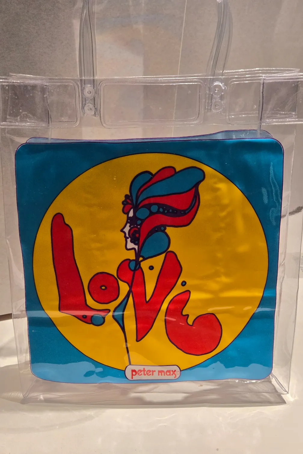 A clear plastic bag in a museum case. On the front of the bag is an image of a woman in a yellow circle against a blue background. The woman’s arms and coat also spell the word LOVE. Peter max is printed at the bottom.