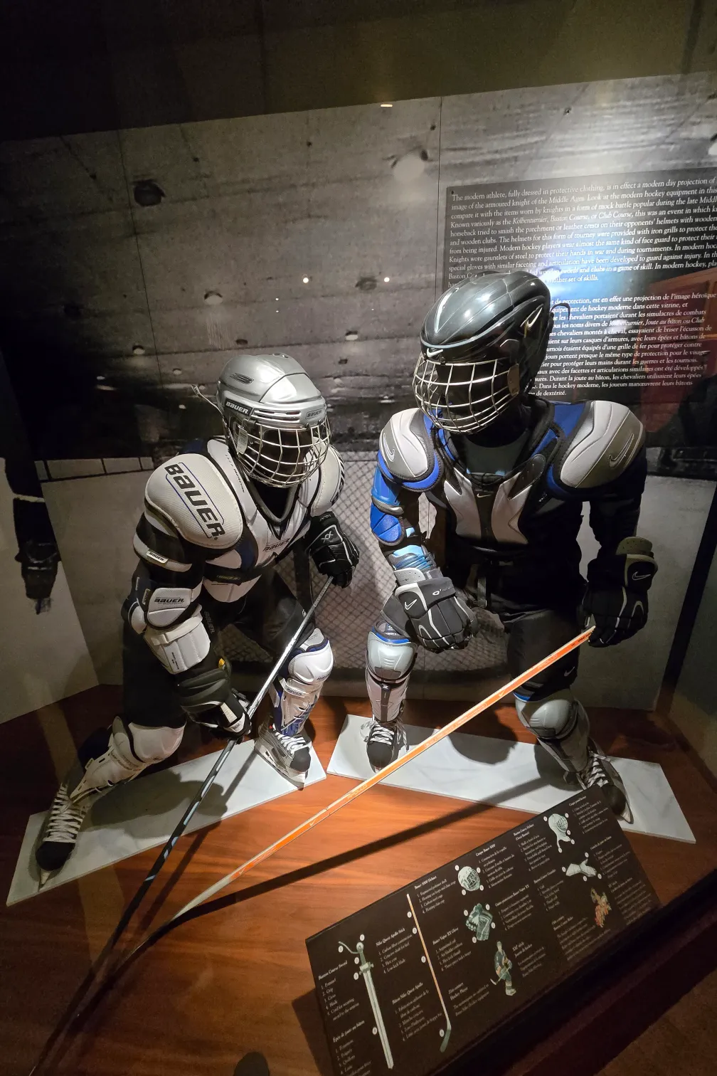 Two mannequins dressed in Bauer hockey gear, including shoulder pads, shin pads, gloves, skates, and helmets. Both mannequins hold hockey sticks and are in dynamic action poses.