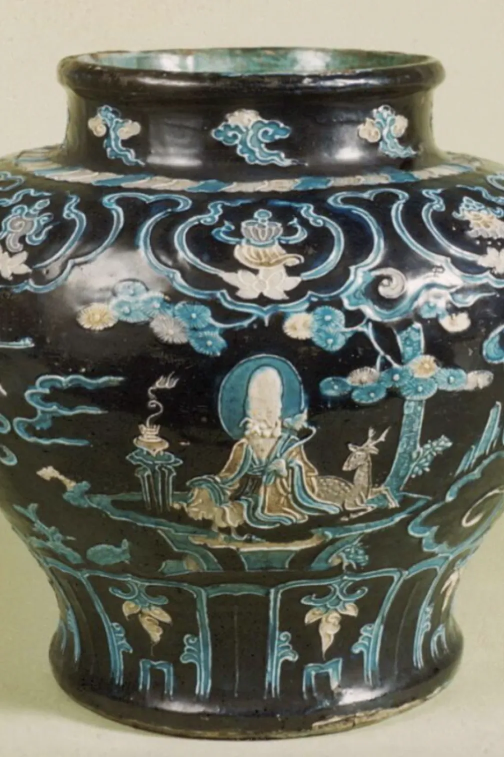 Decorative ceramic jar with a dark, glossy glaze and rounded body. The surface is richly ornamented with raised, multicolored designs in turquoise, blue, white, and pale accents, depicting robed human figures seated or standing in a stylized landscape with clouds, waves, floral motifs, and small animals. Curving bands and framed panels divide the scenes around the vessel, with additional decorative patterns encircling the neck and base. The jar is photographed against a light, neutral background.