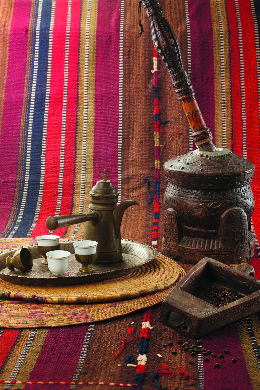 Traditional coffee-serving set with a metal pot and small cups on a tray, alongside carved wooden containers and coffee beans on a colourful striped textile backdrop.