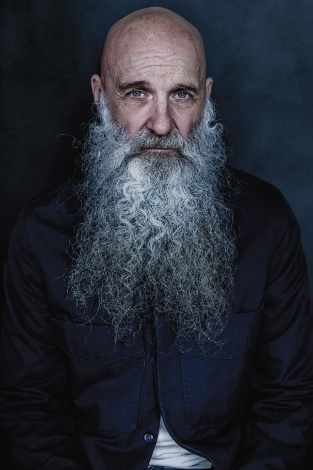 Studio portrait of a bald individual with a long white beard, wearing a dark button-up shirt against a dark blue background.