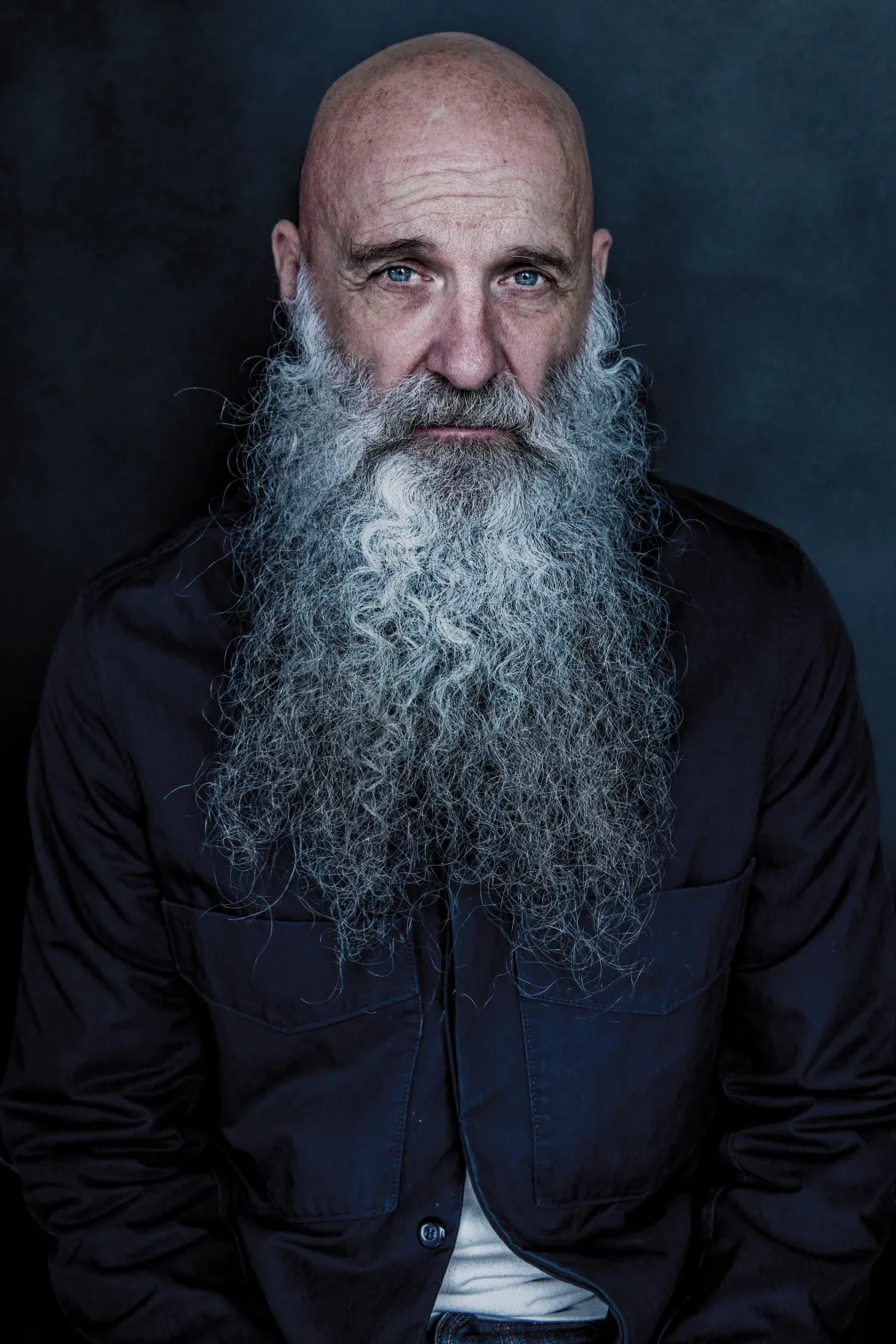 Studio portrait of a bald individual with a long white beard, wearing a dark button-up shirt against a dark blue background.
