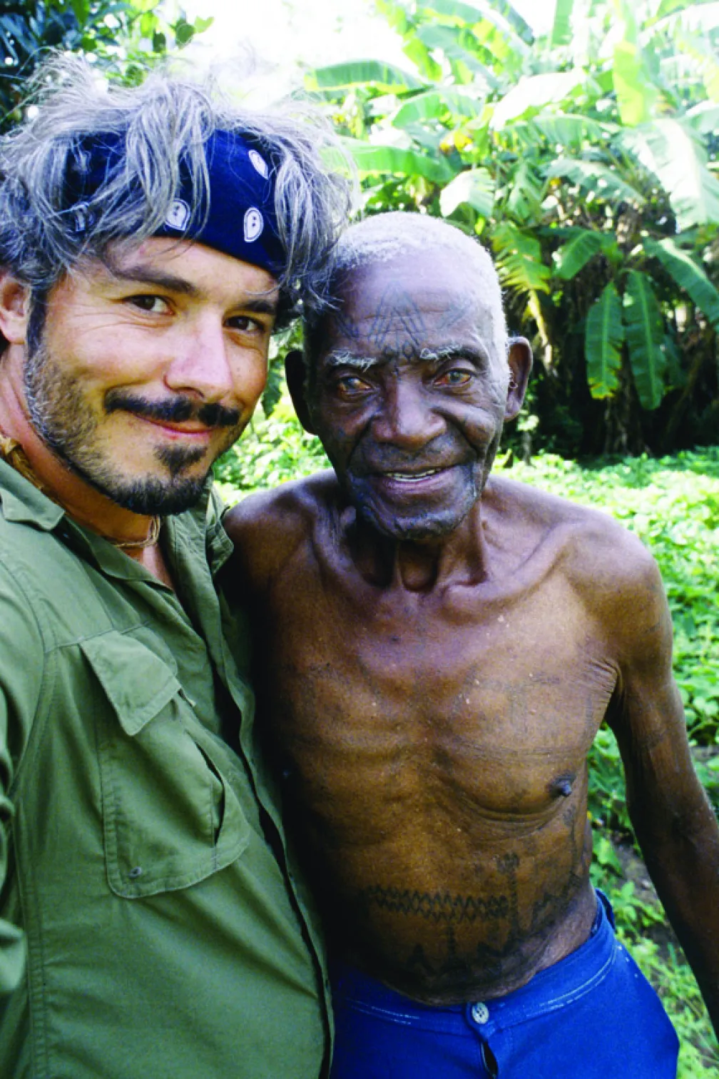 A man in a green shirt and blue bandana taking a selfie with a Makonde tattoo master
