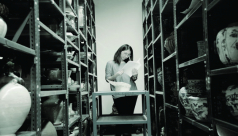 A black and white photograph of a woman standing on a step ladder surrounded by artifacts on shelves 