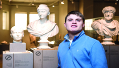 A photograph of a young man in a blue sweater standing in front of three Roman busts.