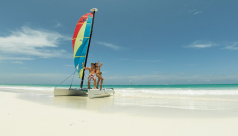 A man & a woman in bathing suits pose on a small catamaran on a sun-lit, white sand beach