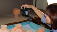 Woman photographing a clay pot, with a cart holding additional pots in the foreground