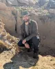 Tom Dudgeson wearing a baseball cap, sunglasses, and dark outdoor clothing crouches beside a large dinosaur skull partially embedded in rocky terrain.