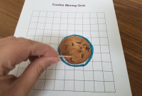 Chocolate chip cookie on grid paper in circle, toothpick poised to begin mining. Hand visible to begin the mining process