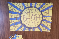 A nearly-complete sun mosaic sits on a table. A dark blue ring and royal blue rays represent the sun. The centre of the sun is filled with yellow. Unused blue and yellow squares sit in a pile next to the mosaic design