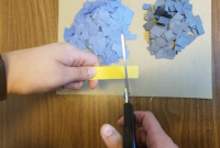 Hands holding scissors cut a yellow strip of paper into small squares onto a tabletop. Piles of pre-cut blue and black squares sit on a piece of paper.