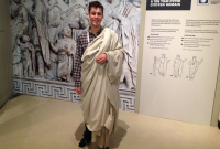 The young man stands with his left arm holding the toga in place.