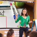 A teacher stands in front of a class. The board behind her shows a paper with a sketch of a sustainable school blueprint, some markers, and a planning sheet. Text says ROM Activity