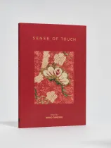 A red hardcover book titled Sense of Touch, edited by Akiko Takesue. The cover features an inset textile artwork with cream, gold, and green embroidered elements on patterned red fabric.