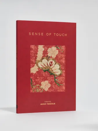 A red hardcover book titled Sense of Touch, edited by Akiko Takesue. The cover features an inset textile artwork with cream, gold, and green embroidered elements on patterned red fabric.