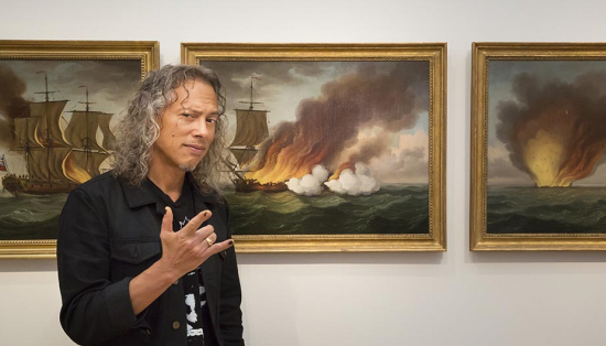 Kirk Hammett standing in front of three paintings of burning ships.