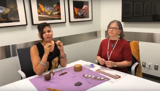 Two women, one with short dark hair and wearing a black dress, and the other with shoulder length graying brown hair and wearing a red top, sit behind a table. The table has several Indigenous ancestor objects on it including a quilled box, a wampum belt, and a spile for tapping maple trees.