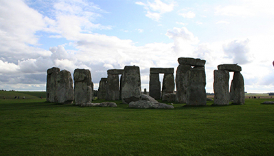 Large rectangular stone pillars in a ring in a field of green grass on a partly cloudy day. Large rectangular rocks placed horizontally on top span between some pairs of pillars. Farm fields are visible in the background.