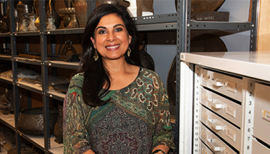 Curator Fahmida Suleman, a smiling woman with tan skin and long dark hair, wearing a colourful print shirt and dangly earrings, stands in front of metal shelving units holding metal Islamic urns and containers.