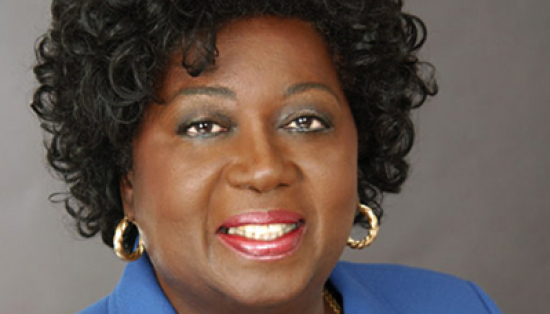Portrait of the Honourable Jean Augustine. She is a smiling middle-aged Black woman with short black hair in loose curls. She is wearing a blue blazer and gold necklace and earrings.