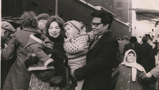 The Lim family stands in a crowded market. Mr. Lim is holding their child in a winter sweater and hat, and Mrs. Lim holds a small monkey wearing pants, booties, and a hooded parka. 
