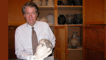 Un homme portant une chemise blanche et une cravate, tenant dans ses mains une tête sculptée. Il se tient devant une armoire remplie de poteries.