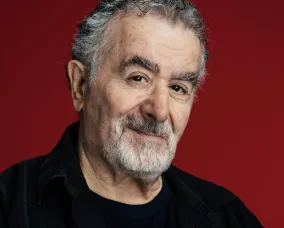 Portrait of an older man with gray hair and a beard, wearing a black shirt and jacket, against a red background.