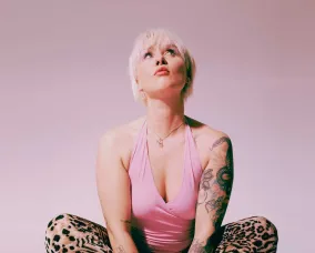 A blonde woman sitting with camouflage pants and a pink top. She is in front of blurred background that is pink and white.