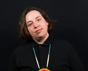 A man posing in front of a camera with a circular necklace in front of a black background.