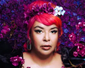 A lady with pink hair and a black top in front of a background filled with purple and blue flowers.