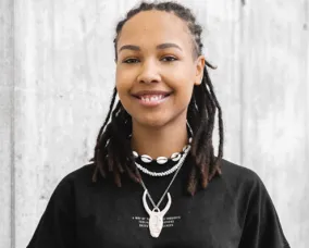 A black woman with braided hair tied back smiles at the camera. She is wearing a black top and white jewellery, including a cowrie shell necklace.