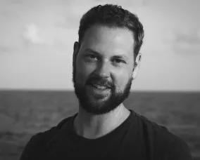 A smiling Caucasian man with dark hair and beard in a black t-shirt.