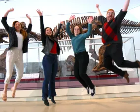 Four adults smiling and jumping in the air in front of a dinosaur skeleton.
