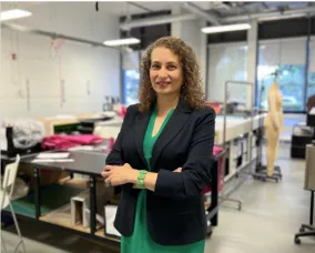 A smiling woman standing and smiling with arms crossed. Behind her is a workroom surrounded by large windows, tables, fabric, a chair and fabric form.   