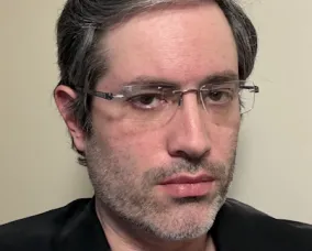 Portrait of Jeffrey Sax with short, dark hair and glasses wearing a black jacket and maroon shirt against a beige background.