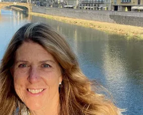 A woman with blonde hair standing beside a river with a bridge in the background.